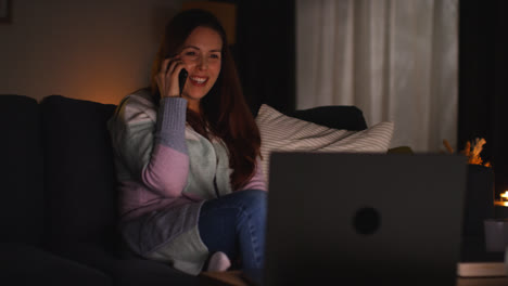 Mujer-Sonriente-Sentada-En-Un-Sofá-En-Casa-Por-La-Noche-Hablando-Por-Teléfono-Móvil-Y-Viendo-Una-Película-O-Un-Programa-En-Una-Computadora-Portátil